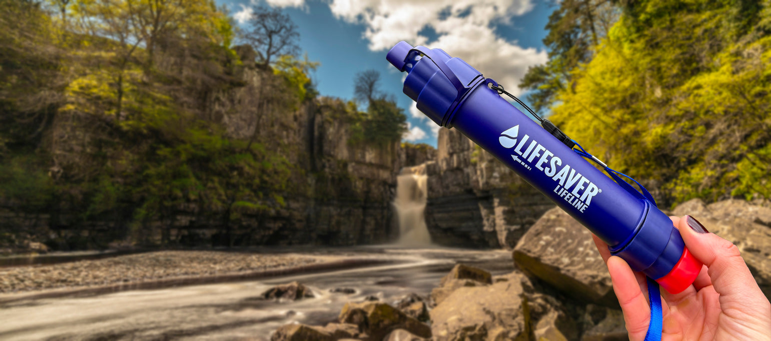 A hand holds a blue Lifesaver Water Filter straw in the foreground, with a blurred outdoor canyon and waterfall landscape in the background.