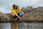 Person in a yellow jacket collecting water from a stream in a rocky landscape
