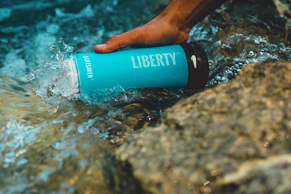 Load image into Gallery viewer, Blue Liberty bottle being submerged in water with a hand holding it.
