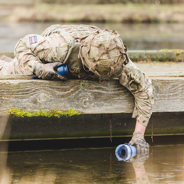 Load image into Gallery viewer, Person in military camouflage lying on a wooden dock with a LifeSaver Bottle, surrounded by water and greenery.
