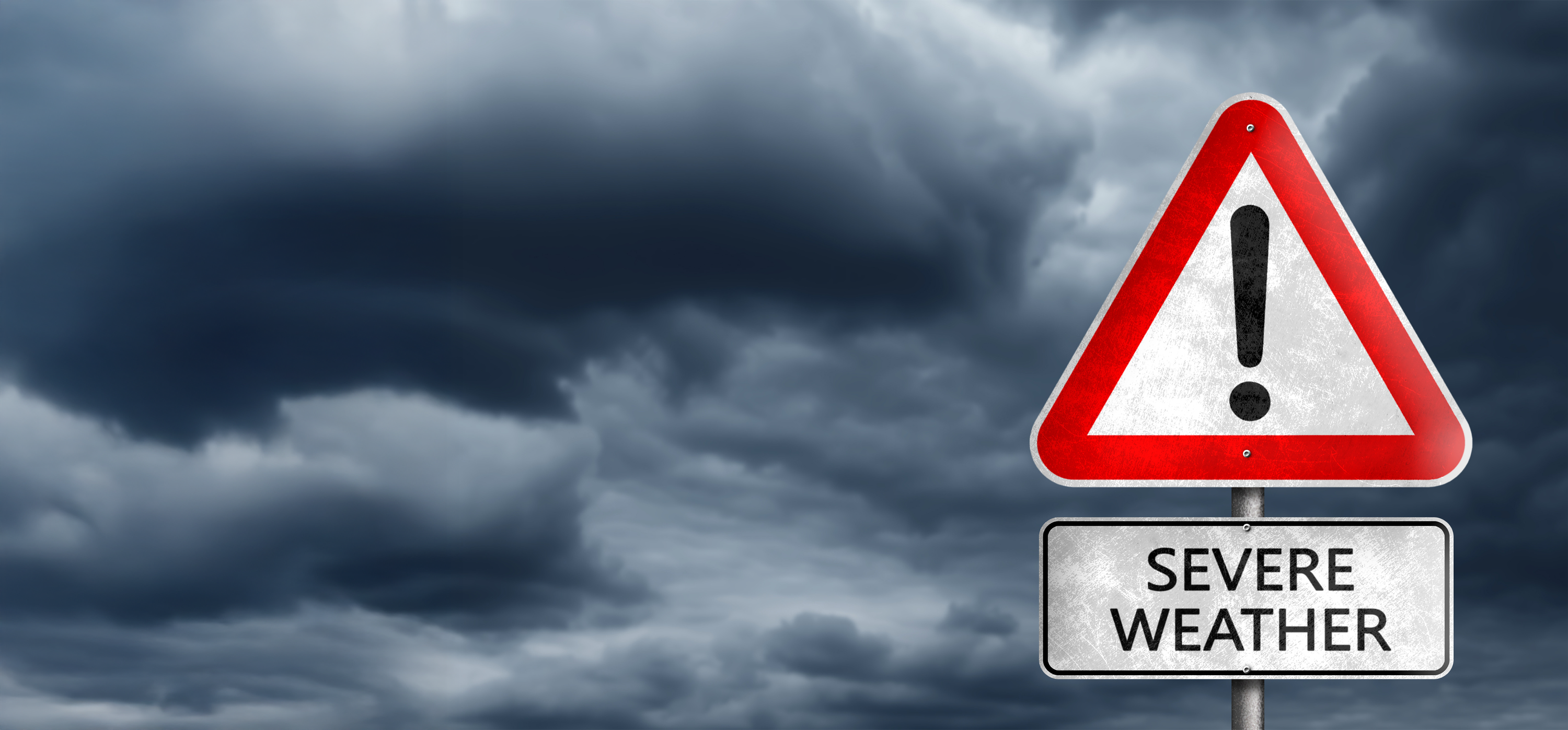 A warning sign with a red triangle and exclamation mark above a white rectangular sign reading 'SEVERE WEATHER,' set against a dramatic dark stormy sky.